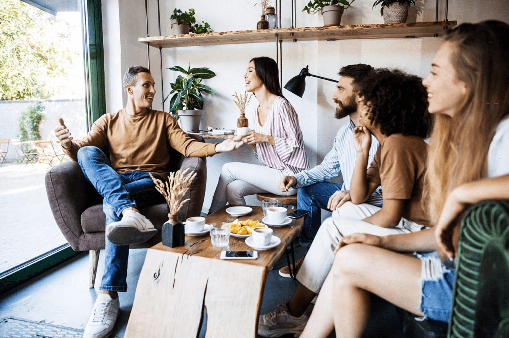 A group of five friends engaged in lively conversation at a cozy café, surrounded by plants, coffee, and snacks, creating a warm and inviting atmosphere.